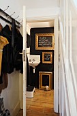 Wandgarderobe im Vorraum und Blick durch offene Tür auf schwarze Wand mit goldenen Bilderrahmen und Eckwaschbecken