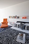 Minimalist bedroom with black and white patterned bedspread, long bookshelf in niche and orange designer swivel chair