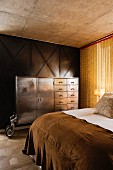 Blick über Bett auf Vintage Metallschrank mit Rollen vor Holzpaneelwand und Kettenvorhang in Schlafzimmer