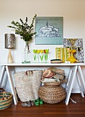 Simple console table with cheerful artworks, vase of flowers and colourful ornaments; various baskets, hats and children's shoes under table