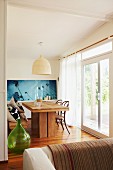 Heavy wooden table and vintage bistro chairs in front of blue photograph in light-flooded dining area; large wicker lampshade above table