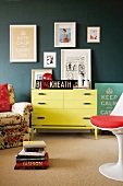 Gelbe Kommode an grauer Wand mit daran hängender Bildersammlung; davor ein Polstersessel mit Blumenmotiv und ein weisser Designer-Drehstuhl mit rotem Sitzkissen