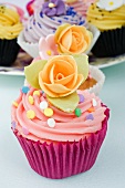 still life of a row of coloured cup cakes decorated with orange rose flowers on top in their cake papers on a white table with other cup cakes in the background