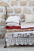 Lace cloth under stacked cushions and blankets on daybed with rusty metal frame against stone wall