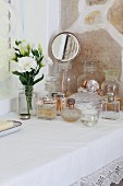 Simple table with white lace cloth and collection of vintage perfume bottles next to vase of flowers and hand mirror in front of stone wall