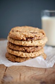 Chocolate chip, peanut butter and oatmeal cookies stacked on baking parchment wih a glass of milk in the background.