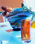 Zylinderförmiges Glas vor Glasvase mit Fackellilien und Rispenzweigen auf Glastisch
