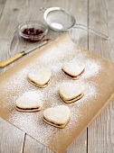 Heart-shaped biscuits filled with jam and dusted with icing sugar on baking paper