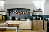 Antique kitchen table with chopping board on table in front of vintage range and collection of copper pans hung in masonry extractor