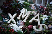 Weihnachtsgesteck aus Tannenzweigen, Scheinbeeren, Lorbeer, Äpfeln, Misteln, Hyazinthen und Pinienzapfen mit Windlicht und Buchstaben XMAS
