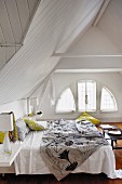 Unmade double bed in charming attic room with segmented arched window and white wooden panelling