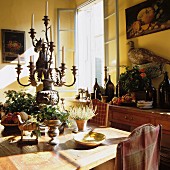 Rustic dining area with magnificent candelabra and fruit and wine bottles on sideboard; still-life paintings and stuffed pheasant on walls