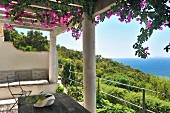Blühende Kletterpflanze auf Pergola einer mediterranen Terrasse und traumhafter Blick über grüne Waldlandschaft und das Meer