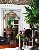 Flowers in a vase on a table across from a highly ornamental wall mirror with a reflection