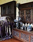 Antique fan and cameras on a carved wooden bench with high back next to a bed with a dark wood headboard and shimmering, violet bed coverlet