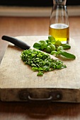 Chopped basil with a knife on a chopping board; a bottle of oil to the rear