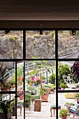 View through open door in floor-to-ceiling, industrial glazing onto platform-style terrace with chairs and potted plants