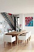 White, upholstered chairs around modern, wooden dining table opposite contemporary photo collage on wall of open-plan interior with staircase