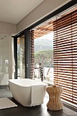 Modern, free-standing bathtub and rattan side table next to glass wall with part-closed wooden louver blinds