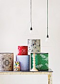 Assorted wallpapered lamp shades on a white, wood table under a hanging light with wire frames