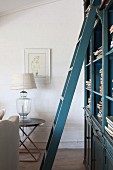 Blue, lacquer ladder in front of a bookcase and table lamp with earth tone lamp shade on a modern side table by the wall