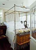 View through open door of four-poster bed with metal frame; vases of flowers on antique chest of drawers at foot of bed