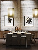 Simple pendant lamps above set table with tablecloth surrounded by barstools in front of whitewashed brick wall in industrial interior