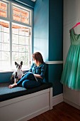 Woman and dog on upholstered window seat against blue-painted wall