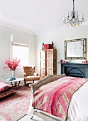 Pink patterned bedspread on antique-style bed in elegant bedroom with upholstered armchair and painted wardrobe in background