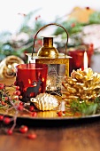 Red glass tealight holder, gilt lantern and pine cone decorations on platter