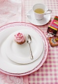 A mini cupcake on a plate with a spoon, and a cup of tea on a checked tablecloth