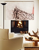 Modernes Gemälde über brennendem Feuer im offenen Kamin; davor ein gemütlicher, weisser Lesesessel