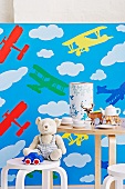 Child's chair and table with toys, child's crockery and money box against wallpaper with aeroplane motif