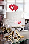 A two-tier wedding cake with red hearts and red writing on an unusual cake stand