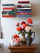 Bouquets of roses in metal jugs and swan-shaped china vase on half-height cabinet below stacked books on minimalist, floating bookshelves