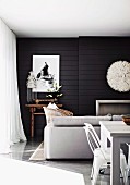 Modern living-dining area in white in front of black wood-panelled wall with white artworks above fireplace and small, antique table