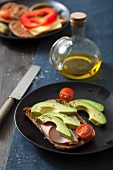 A slice of bread topped with ham, with avocado and tomatoes, and a slice of bread topped with cheese and peppers