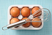An egg whisk and six brown eggs in a porcelain egg box