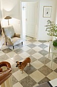 Blick von der Treppe auf Hund im Hausflur mit Schachbrettmustersteinboden grau-weiß und Polstersessel mit Stehleuchte in Zimmerecke