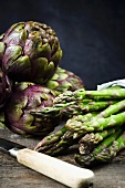 Purple artichokes and stalks of green asparagus on a wooden board