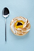 An individual lemon meringue tart and a spoon on a blue surface