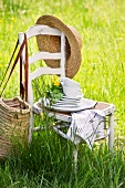 Gestapelte Teller und Tassen mit Blumen auf Holzstuhl in einer Frühlingswiese, Strohhut und Tasche hängen auf der Stuhllehne