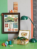 Framed vintage sewing supplies by a wall with a wide stripe; underneath several spools of thread and colorful ribbons in an old cigar box on a green lacquered table