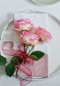 Three pink roses tied with ribbon on a cloth serviette on a charger