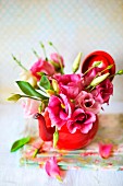 Delicate, pink posy arranged in red teapot; several petals lying on folded floral napkin on white surface