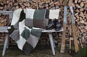 Vintage Skiausrüstung, gestrickte Patchwork Decke und Kissen auf verwitterter Holzbank vor Brennholzstapel