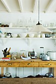 Simple pendant lamp in front of kitchen utensils and appliances on rustic wooden sideboard below bowls and glasses on wall-mounted shelves