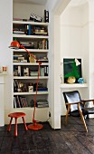 Red, metal standard lamp and red stool in front of fitted bookshelves next to open doorway and view of vintage armchair