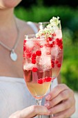 A woman holding two glasses of elderflower fizz with redcurrants