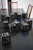 Mehrere Buchstabenwürfel auf dunkler Tischplatte mit Stühlen im Fifitiesstil
